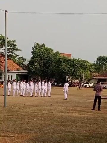 The video shows an outdoor scene on a grassy field with buildings and trees in the background. Several people, mostly dressed in white uniforms, are standing in formation, appearing to be participating in a parade or ceremony. One person in a white uniform is walking and gesturing towards the group. Another person in a dark shirt and pants stands on the right side of the frame, facing away from the camera. The sky is overcast. The video appears to be a short recording of a formal event.