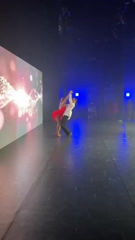 The video captures a dance performance on a stage. Two dancers, a male and a female, are in the center of the stage, engaged in an intricate dance. The male dancer wears a white shirt and black pants, while the female dancer is dressed in a vibrant red, fringed dance dress. They are performing in front of a large screen displaying a luminous, abstract pattern of lights and particles, with a bright white glow at its center. The stage is dimly lit, with blue and purple spotlights illuminating the dancers and the surrounding area. Other individuals are visible in the background, seemingly spectators or other performers, further back on the stage. The floor of the stage appears to be dark and reflective, catching the stage lighting.