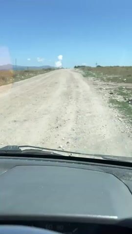 The video is shot from the perspective of a person driving a vehicle. The road ahead is a dirt road that appears to be uphill. The landscape on either side of the road is dry and sparsely vegetated, with some rolling hills in the distance. The sky is clear and blue with a few scattered white clouds. The interior of the vehicle is partially visible, including the dashboard and a steering wheel. A hand briefly appears on the steering wheel, suggesting the driver is making a slight adjustment. The video appears to be taken during daylight hours, likely in a rural or undeveloped area.