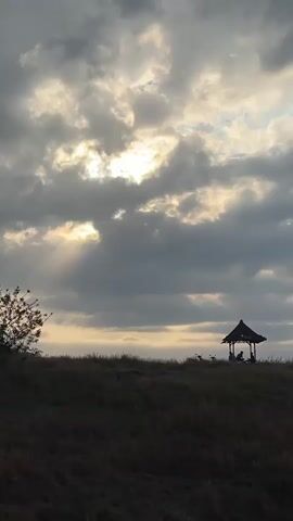 A vertical shot shows a grassy hill with a small pavilion in the distance. The sky is filled with dramatic clouds, with the sun breaking through in a few spots, casting rays of light. The pavilion appears to have at least two people sitting inside. A lone tree is visible on the left side of the frame. The overall impression is that of a serene and possibly remote location.