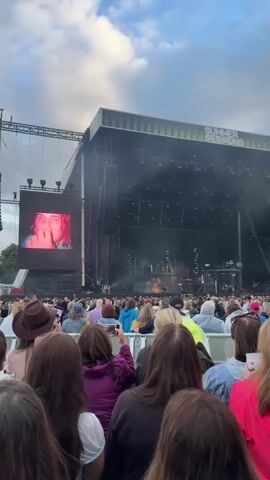 The video shows a large crowd of people facing a stage at an outdoor concert. A large screen on the left side of the stage displays various close-up images of a person's face, which change throughout the video. The stage itself is large and equipped with lighting and sound equipment, with some smoke or haze visible in front of it. The audience is diverse, with people of various ages and genders, many holding up their phones to record the performance. The overall atmosphere appears to be energetic and engaging. The sky above is cloudy, suggesting it might be late afternoon or evening. The text "SUMMER SESSIONS" is visible on a structure above the stage.