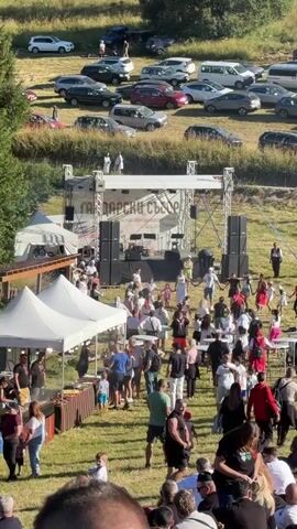 This is a high-angle, wide shot video of an outdoor event. A stage is set up in the middle ground with large speakers on either side. A banner on the stage reads "ГАДАРСКИ СЪБОР". A crowd of people is gathered in front of the stage, many sitting on the grass. Some people are dancing. To the left of the stage are several white tents, possibly for vendors. In the background, a large number of cars are parked on a grassy hillside. The overall impression is that of a festival or fair.