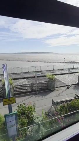 This media file captures a view of the sea at Weston-super-Mare, United Kingdom. The scene is dominated by a wide expanse of water, which appears to be at low tide, revealing a sandy or muddy seabed. Wooden posts are visible in rows extending into the water, possibly marking a path or a former structure.

In the foreground, there is a wooden boardwalk or pier with a glass balustrade. Two large planters, each containing green foliage, are situated on the boardwalk. Beyond the boardwalk, a pathway runs alongside a grassy area with flowering plants and a park bench. A bus stop sign is also visible, with the text "No stopping except local buses" clearly legible on a yellow sign attached to it.

The sky is bright and partly cloudy, suggesting it is daytime, likely mid-morning or afternoon. The overall atmosphere is peaceful and serene. The location cues, such as the distinctive coastline in the distance and the presence of a pier, strongly indicate Weston-super-Mare. No specific activity or event is prominently featured, but the scene conveys a sense of a quiet day by the seaside.