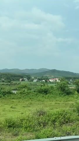 The video, shot from inside a vehicle, shows a landscape with green fields and trees in the foreground. In the distance, there are rolling hills and mountains under a bright sky with scattered clouds. A white sign with Vietnamese writing, presumably advertising a company, is visible in the lower right corner of the frame. The vehicle appears to be moving, providing a slightly shaky perspective of the passing scenery.