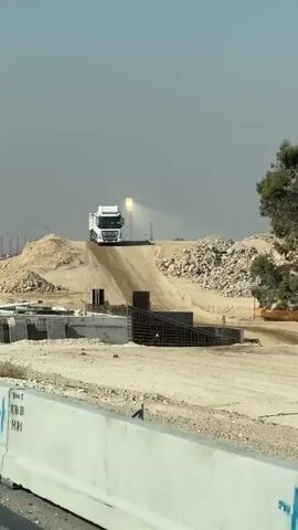 A white dump truck drives up a dirt ramp at a construction site under a hazy sky. A tall wooden pole stands on the left side of the frame. An orange and white striped pole with an orange circular sign is on the right. In the background, construction equipment and infrastructure are visible, including what appears to be a drilling rig.