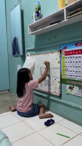 A young girl with long dark hair, wearing a pink t-shirt and a blue skirt, sits on the floor facing a whiteboard. She is drawing or writing on the whiteboard with a marker. To her right, a green pencil and a blue toy car are on the floor. Behind her, the whiteboard is covered with writing and drawings. To the right of the whiteboard, a colorful alphabet chart is taped to the wall. Above the whiteboard, a white shelf holds various items, including a blue spray bottle and books. The wall is painted a light blue-green color, and the floor is tiled with white squares. A mirror is visible on the left side of the image, reflecting a blue door and a hanging garment. The scene appears to be an indoor setting, possibly a child's room or play area.
