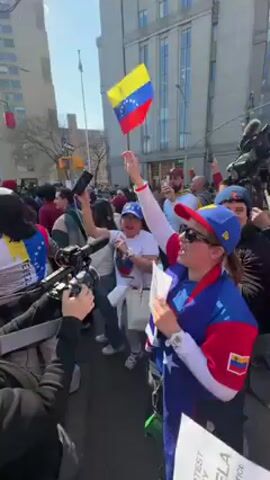 The video captures a vibrant street protest or demonstration, likely in a city environment given the tall buildings in the background. The weather appears sunny and clear. A large crowd of people is gathered, many holding small Venezuelan flags and wearing apparel featuring the flag's colors (yellow, blue, and red with stars).

In the foreground, a woman wearing a blue and red top with a Venezuelan flag draped over her shoulders, a red cap, and sunglasses, is actively participating. She holds a small Venezuelan flag in her right hand and a sign in her left. The sign partially visible reads "PEACEFUL PROTEST FOR DEMOCRACY VENEZUELA" and another phrase that appears to be "NO MORE DICTATORS." She is cheering loudly.

Next to her, another woman in a white t-shirt, white jacket, and a yellow cap is also engaged, holding up a smartphone as if recording or taking pictures. She is also actively cheering.

Throughout the crowd, other individuals are visible, many holding up their phones, suggesting they are capturing the event. Some professional-looking camera equipment is also visible on the left side of the frame, indicating media presence. The overall atmosphere appears enthusiastic and passionate.

The audio features a clear chant from the crowd: "Yo vine porque quise, a mí no me pagaron. Yo vine porque quise, a mí no me pagaron," which translates to "I came because I wanted to, they didn't pay me. I came because I wanted to, they didn't pay me." This chant implies a grassroots, voluntary participation in the protest, without monetary compensation.