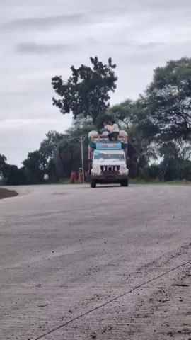 A white Mahindra Bolero pickup truck is driving on a paved road with a dirt shoulder. The truck is loaded with goods, covered by tarps, and has decorative elements on its sides. Two men are sitting on top of the load. The sky is overcast. Trees and some foliage are visible on the sides of the road. In the background, a few people are standing near a pole on the left side of the road. The video has a slightly grainy quality.