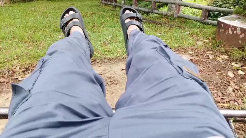 The video shows a first-person perspective of a person seated with their legs extended and swinging back and forth. The person is wearing gray cargo pants and black sandals. The ground below alternates between dirt with leaves and grass. In the background, there is a wooden fence and some greenery. The camera angle is stable, focusing on the person's legs and the immediate ground area.