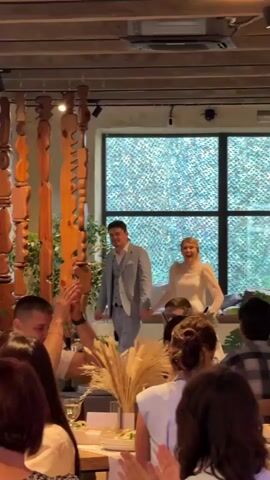 A wedding reception is underway in a dimly lit venue with wooden beams and decorative wooden hanging art. A bride in a white dress and groom in a light blue suit are entering the room, holding hands and smiling. The groom appears to be speaking. A person is filming them with a smartphone from the audience. Other guests are seated at tables.