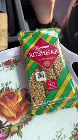 This is a first-person perspective video. The video opens on a close-up of a package of sunflower seed kozinak, which is a type of confection. The package has green and yellow stripes and an image of a sunflower. The camera then pans to the left, revealing a wooden cutting board with a piece of lard on it and a knife. Next to the cutting board is a white mug with dark liquid in it, presumably tea or coffee, with a spoon inside. The camera then moves to the right, showing a purple plastic bowl and a plastic bag filled with what appears to be chopped vegetables. Further to the right, a camping stove with a pot and a kettle are visible. The background shows a blanket with a star pattern and some pillows. A bottle of water is also visible on the left side of the frame. The scene appears to be set up for a meal or a snack, possibly during a camping trip or in a small living space.