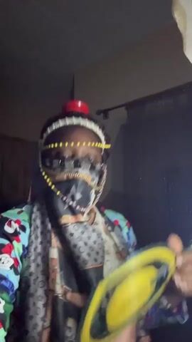 A person wearing a colorful jacket, a scarf with a designer print, a beaded headdress, and a face mask is visible in a dimly lit room. The person is dancing and moving their arms. A window with dark curtains is partially visible in the background. The scene appears to be indoors.