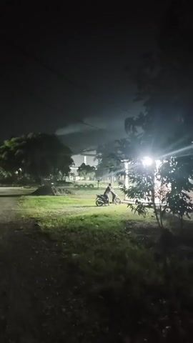 A person is sitting on a motorcycle on the side of a dirt road at night. There are trees and some buildings in the background, illuminated by artificial lights. The scene appears to be an outdoor area with some grass and dirt.