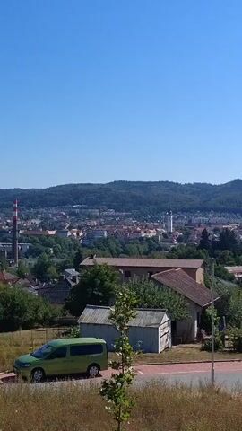 The video shows a panoramic view of a town on a clear sunny day. The foreground features dry grass and a paved pathway. A green van is parked to the left of the pathway. In the middle ground, there are houses with red roofs and trees. Further in the distance, the town is visible with various buildings, including a tall industrial chimney with red and white stripes. A hill covered with trees forms the backdrop of the town. The sky is a clear, bright blue. The camera appears to be panning slowly to the right.
