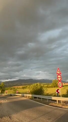 The video captures a scenic driving moment in Barakovo, Bulgaria, during the late afternoon or early evening. A silver Suzuki Jimny is seen driving around a bend on a paved road, which is bordered by a metal guardrail. The sky is predominantly cloudy, with heavy gray clouds, but a patch of golden sunlight illuminates parts of the landscape on the left. On the left side of the road, several European-style road signs are visible: a yellow diamond indicating a priority road, a blue circular sign with opposing white arrows signifying two-way traffic, and a red and white striped hazard marker. As the car navigates the curve, red and white chevron signs marking the sharp bend are visible on the right. The surrounding landscape features vibrant green fields, abundant trees, and distant mountains, some of which appear to have snow-capped peaks or light-colored rock formations. People can be vaguely seen inside the car.