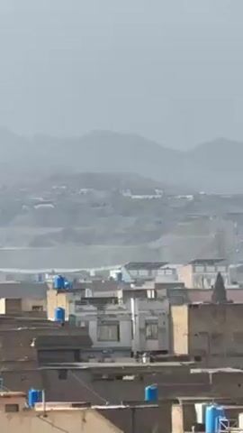 The video shows an overview of a densely populated area with buildings of varying heights, likely a town or city. In the background, there are mountains visible under a hazy sky. Many of the buildings in the foreground have blue water tanks on their roofs. The video then pans out to show a wide, barren field in the foreground under a cloudy sky. The landscape appears dry and sandy.