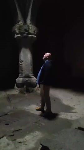 A man with a shaved head wearing a dark jacket and khaki pants stands in a dimly lit, possibly stone structure. He is looking up at a large stone column that flares out at the top into arching supports. The column is weathered and appears quite old. The surrounding area is dark, and the ground is uneven. The man is the only person visible in the frame. He seems to be observing the architecture of the structure.