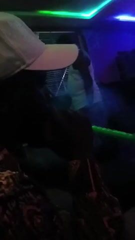 The video shows a person from the chest up, positioned in what appears to be a dimly lit lounge or club environment. The person is wearing a white baseball cap, a dark-colored inner shirt, and a patterned outer shirt. They are actively smoking from a hookah or shisha pipe, holding the mouthpiece to their lips and exhaling large, dense clouds of smoke.

The lighting in the room is characterized by vibrant LED lights. There are green LED strips visible along the ceiling on one side, and blue/purple lighting illuminating another part of the ceiling and wall. These colored lights interact with the exhaled smoke, causing it to appear tinted green, blue, or purple depending on the angle and the light source.

In the background, a window with horizontal blinds is visible. Another person, wearing a white top, can be faintly seen standing behind the blinds, but their features are indistinct due to the low light and the blinds obscuring them. The overall atmosphere seems relaxed and social, possibly enhanced by the background music that can be heard. The person smoking appears calm and focused on the activity.