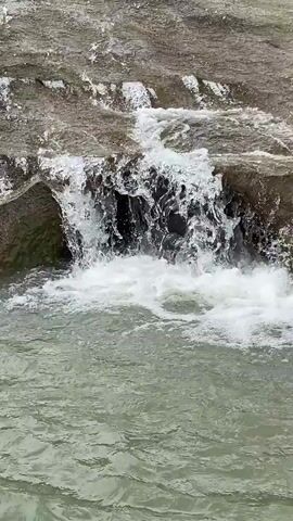 The video shows a small waterfall flowing into a body of water. The waterfall appears to be created by water flowing over rocks. The water is relatively clear and reflects light, and it is creating white foam as it crashes against the rocks. The surrounding environment appears to be natural. The overall scene is a close-up view of the water and the rocks.