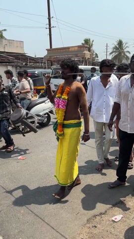 A video shows a religious procession outdoors on a sunny day. People are walking on a paved street with buildings and cars in the background. Some participants have metal rods piercing their cheeks. One man wears a yellow sarong and floral garlands, and raises his hand. A boy, adorned with jewelry and a garland, carries a yellow flag. Another man holds a platter.