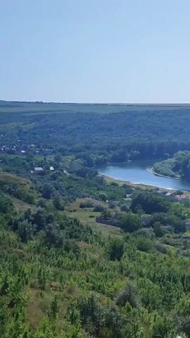 The video is a landscape view of a river, a small village, and lush green hills under a clear blue sky. The river is wide and reflects the sky. The village has scattered houses nestled among trees. The hills in the foreground are covered with dense vegetation and some scattered trees. The background features rolling hills covered in trees, creating a layered effect. The lighting suggests it is daytime, likely late morning or early afternoon. There are no people or animals visible in the video.