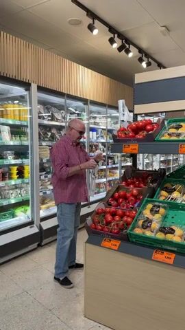 A man in a pink checkered shirt and jeans stands in a grocery store aisle, looking at a plastic bag and what appears to be a receipt or tag. He is positioned in front of a display of tomatoes, with refrigerated cases filled with food products behind him. The store lighting fixtures are visible on the ceiling.