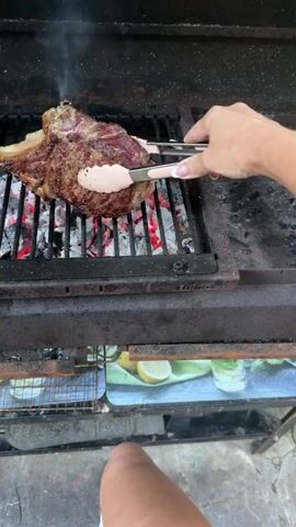 A close-up shot shows a raw steak being grilled over hot coals. A person's hands, with manicured nails, are using white and silver tongs to turn the steak. Smoke is rising from the grill. The steak has a bone protruding from one side. The grill is black and appears to be made of metal. Below the grill, there is a colorful decorative surface with images of lemons and limes. The lower part of the shot shows a person's bare leg.