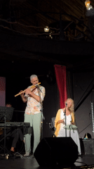 Live indoor set: keys, sax, flute, and percussion on a decorated stage.