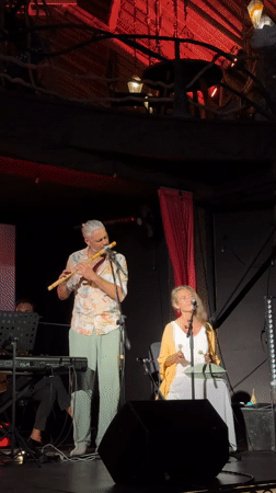 Live indoor set: keys, sax, flute, and percussion on a decorated stage.
