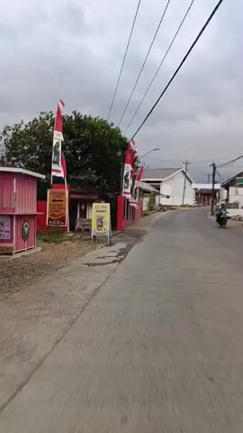 The video begins with a view of a street scene. On the left side of the road, there is a red building with several flags displayed prominently. These flags are white and red, with black and white images of a person on them. There is also a sign with Indonesian writing and an image of a drink. A smaller sign next to it advertises "ES TEH JUMBO". The building appears to be a shop or a business. Further down the street, more of the red building is visible, as well as other structures that appear to be houses. The road is paved and curves to the right. There are power lines overhead. The sky is overcast. On the right side of the street, there are also some houses and vegetation, including banana trees. The video appears to be recorded from a moving vehicle, possibly a motorcycle or bicycle, judging by the slight motion blur and perspective. The overall impression is a street in a residential or commercial area, possibly in Southeast Asia, given the flags and writing.