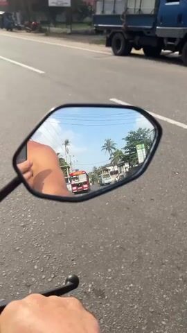 The video is a first-person perspective from a motorcycle, focusing on the reflection in the right-side mirror. The mirror shows a street scene with a red and white bus approaching from a distance. To the left of the bus, a green and yellow tuk-tuk is visible, and further left, there are buildings and palm trees. In the foreground of the mirror's reflection, a bare leg and arm are visible, indicating the rider is wearing shorts and possibly has their arm extended. The road surface is asphalt, and the weather appears to be sunny with a clear blue sky. Other vehicles, including cars and motorcycles, can be seen on the road in the background of the reflection and in the out-of-focus foreground.