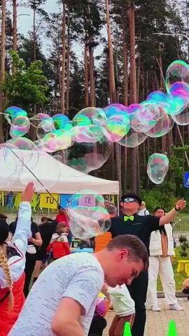 A man in a black shirt and green bow tie is creating giant bubbles in an outdoor setting. He holds two sticks with a string between them, dipping the string into a bubble solution and then spreading the sticks apart to form large, iridescent bubbles that float into the air. Several people, including children, are in the background, observing the activity. A few tents are visible, and tall trees line the back of the scene. The weather appears to be overcast.