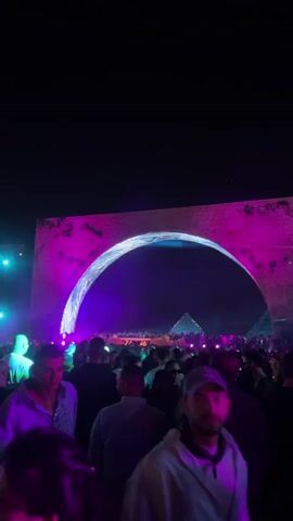The scene shows a crowded music festival at night. In the foreground are many people, mostly men, standing and looking toward the stage. The stage is framed by a large stone-like archway, which is lit with purple and blue lights. Through the arch, a pyramid-shaped structure is visible, also illuminated with colored lights. The words "ZAMNA" are displayed on the stage. The overall atmosphere suggests a lively and energetic event, with music and light creating a vibrant spectacle.
