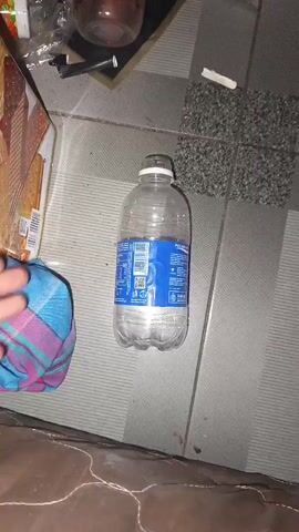 The video pans across a floor with grey and white tiles. On the floor are several objects: a clear plastic water bottle with a blue label, a cigarette, a box of crackers, and other unidentifiable items. A hand with a colorful cloth is visible in the lower left corner of the frame. The lighting suggests it is nighttime or indoors with artificial light.