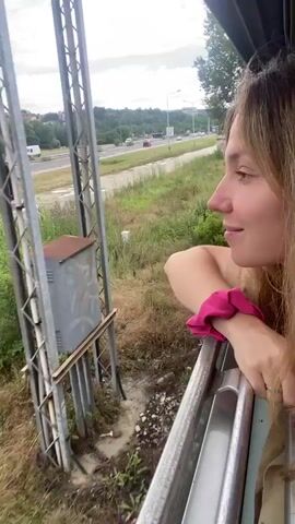 A woman leans out of a train window, her arm resting on the window frame. She has blonde hair and is smiling. A pink scrunchie is visible on her wrist. Outside the window, a highway can be seen with several black vans driving on it. There is also lush green vegetation and trees. A stop sign is visible by the side of the road. The lighting suggests it is daytime.