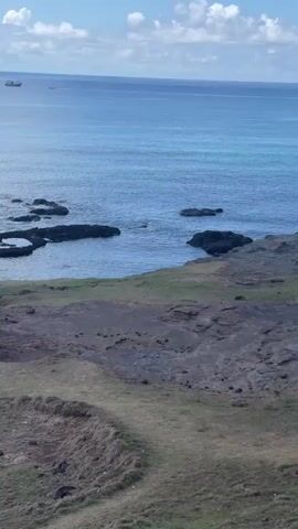Here's a description of the video:

The video shows a view of the ocean from a rocky shoreline. The sea is calm, with a boat visible in the distance. The foreground consists of a grassy slope that descends to a rocky area along the water's edge. Two people are present: one initially walks along the shoreline, then stops and appears to throw a rock into the water. The second person is standing and observing. After the rock is thrown, the camera pans to the left and zooms in slightly, shifting the focus away from the two people toward a small strip of beach.