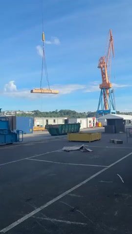 The video shows a lift operation underway at what appears to be a port or industrial site. A crane is lifting a rectangular wooden object with straps. The camera moves from a wide shot, showing the crane, surrounding containers, and temporary buildings, to a view directly below a blue metal structure of the crane, looking up at the wooden object suspended by the crane's cable against a clear blue sky. A smaller orange crane stands nearby. A worker in a high-visibility vest walks in the background. The ground is asphalt, marked with white lines, suggesting a parking or storage area.