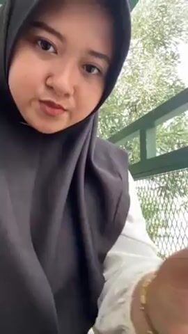 A person wearing a black hijab and white shirt is holding up a tall glass of iced coffee. The drink has a light yellow layer at the bottom, a dark brown layer above, and ice cubes at the top. The person stirs the drink with a black straw. The background shows a green metal structure and trees.