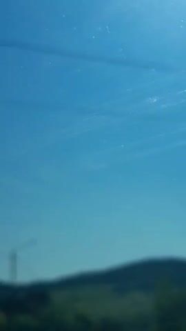 The video shows a view through a dirty glass surface. The sky is a bright, clear blue with faint, hazy white streaks across it, possibly from dust or reflections on the glass. The bottom portion of the video is a blurry, dark green and black landscape, suggesting trees and hills. The focus is on the sky, with the landscape appearing out of focus and distant. There are no discernible people, objects, or actions taking place.
