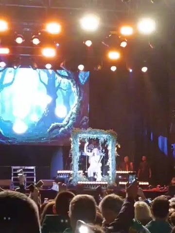 The video shows a live concert performance. A singer in a white outfit sits on a swing decorated with greenery, positioned in front of a large screen displaying an animated forest scene with glowing orbs. The stage is lit with bright, warm lights. Behind the singer are musicians playing instruments, and in front of the stage, a crowd of people is visible, many holding up their phones to record the performance.