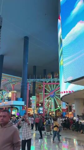 The video captures a vibrant indoor theme park crowded with people. The camera pans from right to left, revealing a large screen, a rollercoaster track, and a Ferris wheel. Many visitors are walking around, some taking pictures. Overhead, there are suspended speaker systems and several large pillars supporting the ceiling. The overall atmosphere is bustling and colorful.