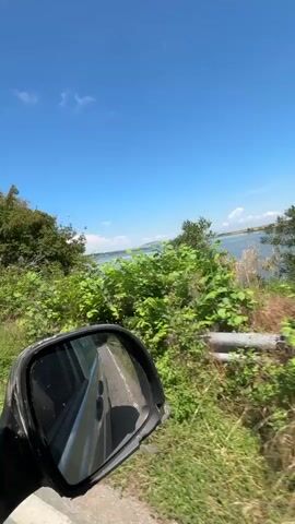 This is a first-person perspective video taken from inside a vehicle, likely a car, driving along a road. The camera is positioned to capture the view through the side window and the side-view mirror. The exterior scene features lush green vegetation, trees, and a body of water in the distance, under a bright blue sky with some clouds. The side-view mirror reflects the road behind the vehicle, showing a white lane marker and other cars in the distance. The overall impression is of a scenic drive through a natural landscape.