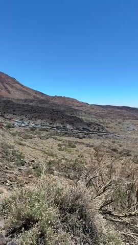 A drone shot slowly pans across a vast, arid landscape under a clear blue sky. The terrain is rugged and volcanic, with rocky slopes covered in sparse, dry vegetation. A winding road snakes through the scene, with a few cars visible in the distance. The landscape is dominated by the imposing presence of a large volcano in the background. The overall impression is one of a remote, desolate, and natural environment.