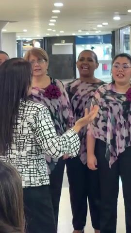 A small choir of women, all wearing similar black pants and lavender-patterned tunics, is rehearsing or performing in what appears to be a shopping mall. The woman facing them, presumably the conductor, has long dark hair and wears a black and white patterned jacket, using her hands to lead the group. The singers are of diverse ages and ethnicities. The mall setting is indicated by the store fronts and ceiling lights visible in the background. A few bystanders can be seen watching the performance.