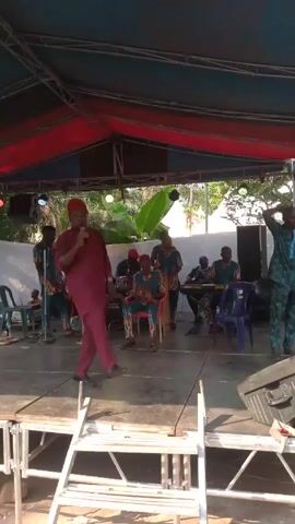 A band is playing on an outdoor stage. The stage is covered with a metal frame and has a blue and red tarp overhead. A man in a burgundy suit and matching hat is standing in front of a microphone, singing and dancing. Behind him, five other men are playing instruments: drums, keyboard, and stringed instruments that are being plucked. They are all wearing matching blue and orange patterned shirts. There are speakers and chairs on the stage. Behind the stage, there is a white cloth backdrop and trees. Some people are sitting or standing near the stage.