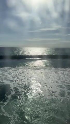 The video shows an ocean scene with waves and the sun reflecting on the water. The sun's rays are visible, creating a bright glare. The water in the foreground is foamy and agitated, indicating movement. The sky is partly cloudy. There are no people or man-made objects visible. The overall impression is of a natural, dynamic seascape.