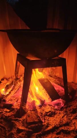 The video shows a black cauldron sitting on a black metal stand over a burning wood fire. The fire is contained in a sandy area with an orange metal wall in the background. A hand reaches in to add more wood to the fire. The overall lighting is dim, with the fire providing the main source of light. The video appears to be taken indoors, in a contained area.