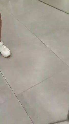 The video shows a woman in a yellow off-the-shoulder top and white skirt taking a selfie in a room with mirrored walls and ceiling. She is wearing a white baseball cap and white sneakers. The mirrors create a kaleidoscopic effect, showing multiple reflections of the woman and the room, which is decorated with hanging white and green floral arrangements. The floor is a light-colored tile. In the first few seconds, a close-up of the woman's lower legs and feet in white sneakers is visible. The rest of the video focuses on the woman and her reflections, as she moves slightly and adjusts her phone while taking pictures.