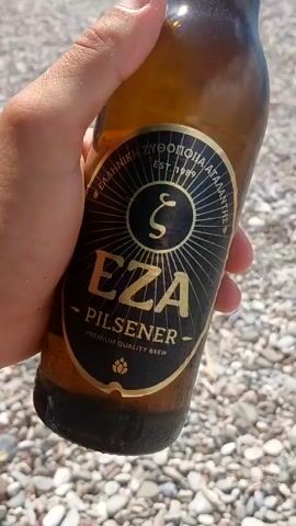 A person's hand holds a brown glass bottle of EZA Pilsener beer on a pebbled beach. The bottle label is visible and shows the brand name, type of beer, and "EST. 1989". The background is a body of water with a hazy shoreline in the distance under a blue sky with white clouds.