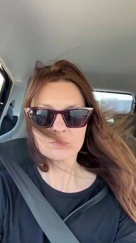 A woman with long, reddish-brown hair, wearing a black t-shirt and sunglasses, is seated in the passenger seat of a car. Her hair is being blown around by the wind. She is looking directly at the camera and her lips are pursed. The seatbelt of the car is visible across her chest. The interior of the car is dimly lit, with the outside visible through the windows, showing trees and a clear sky.